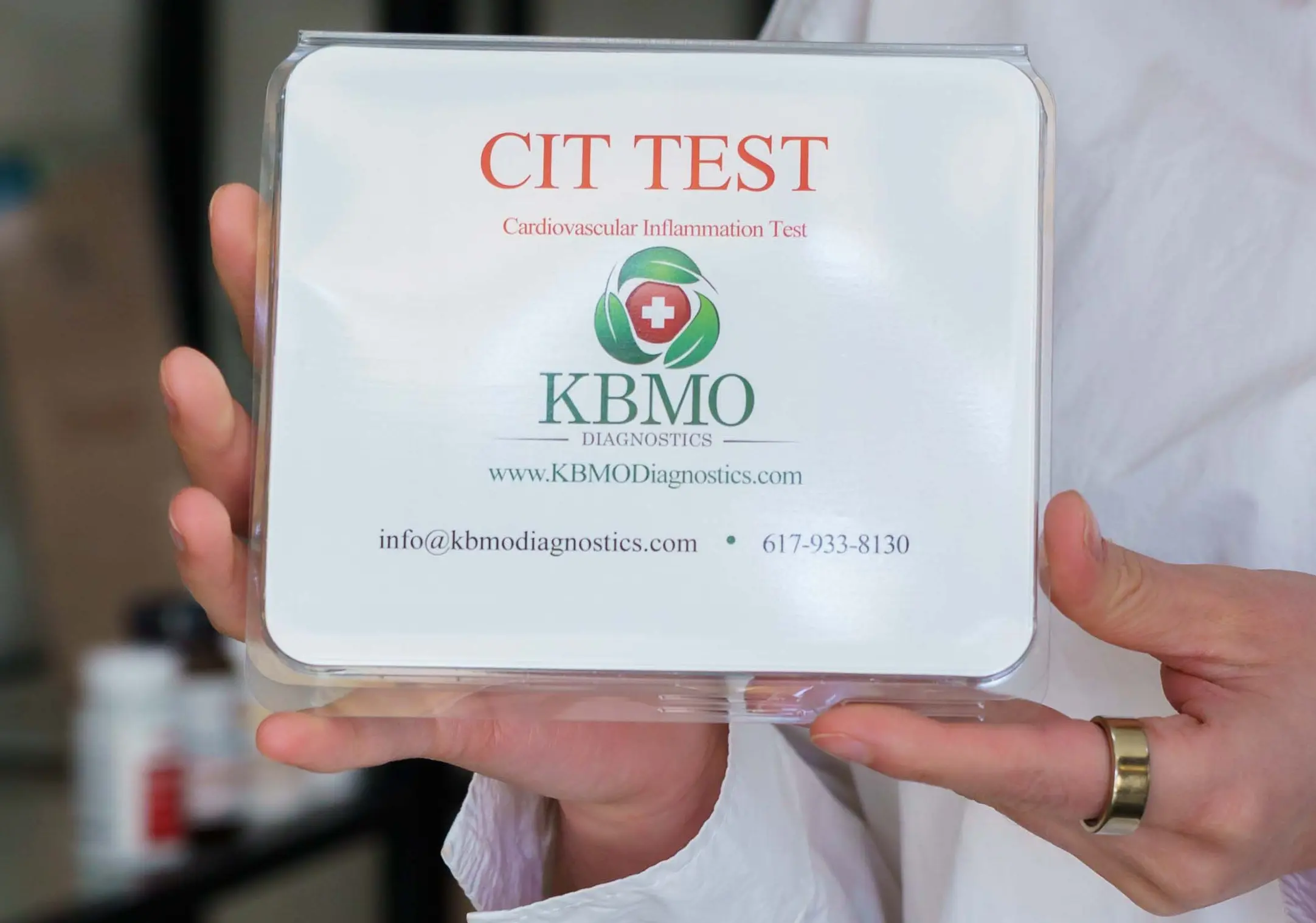 A hand holds a CIT TEST box labeled KBMO with contact details.